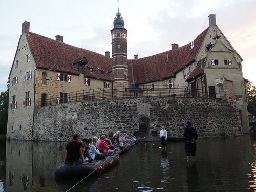 Zuschauer sitzen in Schlauchbooten auf dem Wasser vor der Burg Vischering, während Schauspieler barfuß im Wasser eine Szene aufführen.