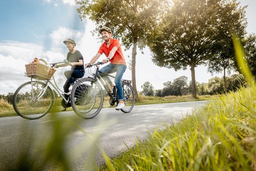 Zwei Erwachsene fahren nebeneinander auf Fahrrädern auf einer ruhigen Landstraße. Beide tragen Fahrradhelme und lächeln. Die Frau links hat einen Korb mit einem orangefarbenen Tuch auf dem Lenker ihres weißen Fahrrads. Der Mann rechts trägt ein rotes Poloshirt und fährt ein schwarzes Fahrrad. Die Sonne scheint durch die Bäume am Straßenrand, und die Szene wirkt freundlich und sommerlich. Im Vordergrund ist grünes Gras zu sehen, im Hintergrund Bäume und ein blauer Himmel mit einigen Wolken.