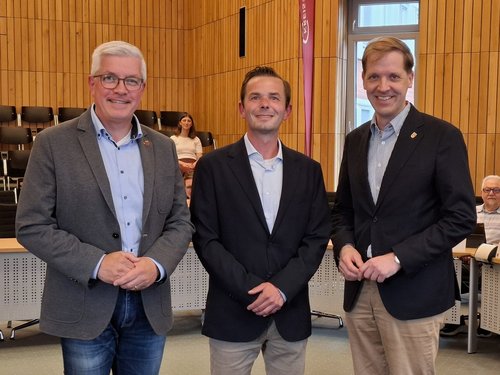 In einem Sitzungssaal stehen drei Männer mit Jackett nebeneinander. Sie haben die Hände verschränkt und lächeln in die Kamera.