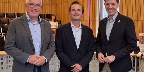 In einem Sitzungssaal stehen drei Männer mit Jackett nebeneinander. Sie haben die Hände verschränkt und lächeln in die Kamera.
