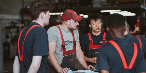 Vier Männer stehen im Kreis um einen roten gegenstand herum. Alle tragen rot-schwarze Latzhosen. Drei von Ihnen tragen ein schwarzes T-Shirt darunter.