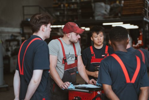 Vier Männer stehen im Kreis um einen roten gegenstand herum. Alle tragen rot-schwarze Latzhosen. Drei von Ihnen tragen ein schwarzes T-Shirt darunter.