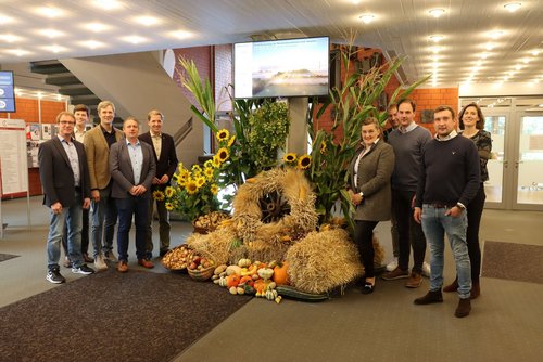 Zehn Personen stehen vor einem geschmückten Ernterad in einem Foyer.
