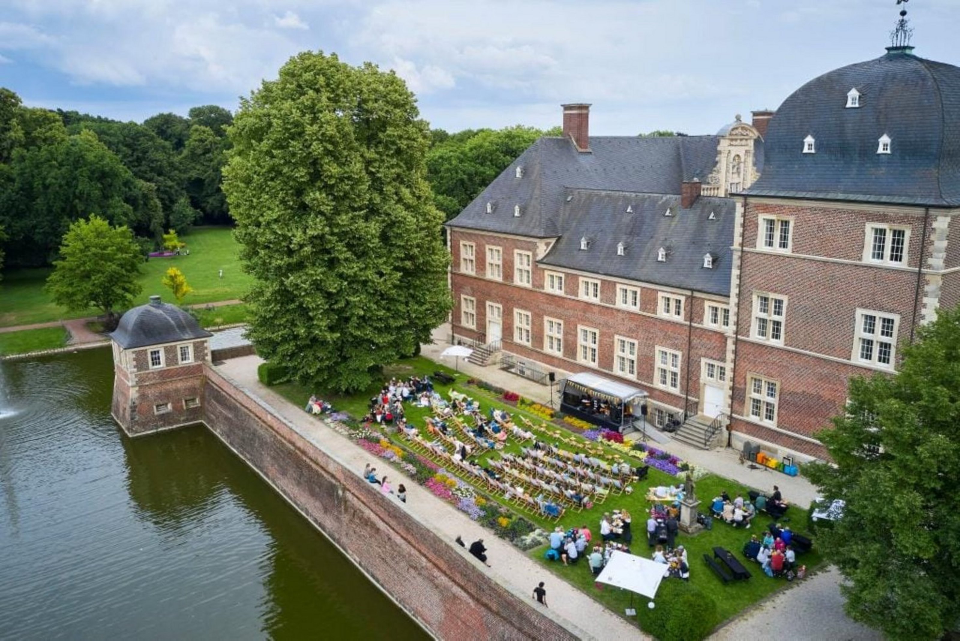 Luftaufnahme eines Schlosses mit Wassergraben, vor dem viele Besucherinnen und Besucher bei einer Veranstaltung im Schlossgarten sitzen.