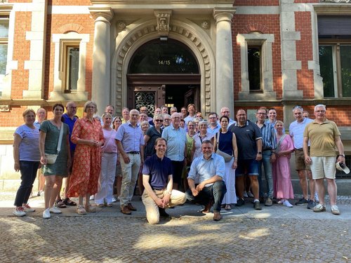 Vor der Eingangstür eines historischen Backsteingebäudes steht eine Gruppe von 38 Personen in mehreren Reihen. In der Mitte knien zwei Männer. 
