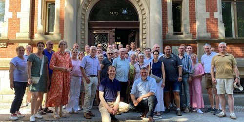 Vor der Eingangstür eines historischen Backsteingebäudes steht eine Gruppe von 38 Personen in mehreren Reihen. In der Mitte knien zwei Männer. 