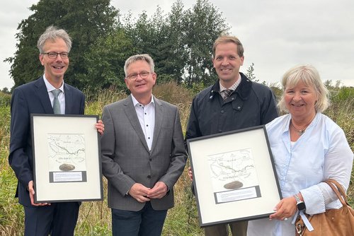 Vier Personen stehen in der Natur auf einer Wiese. Im Hintergrund ist ein Gebüsch. Zwei der Personen halten eine Urkunde in der hand.