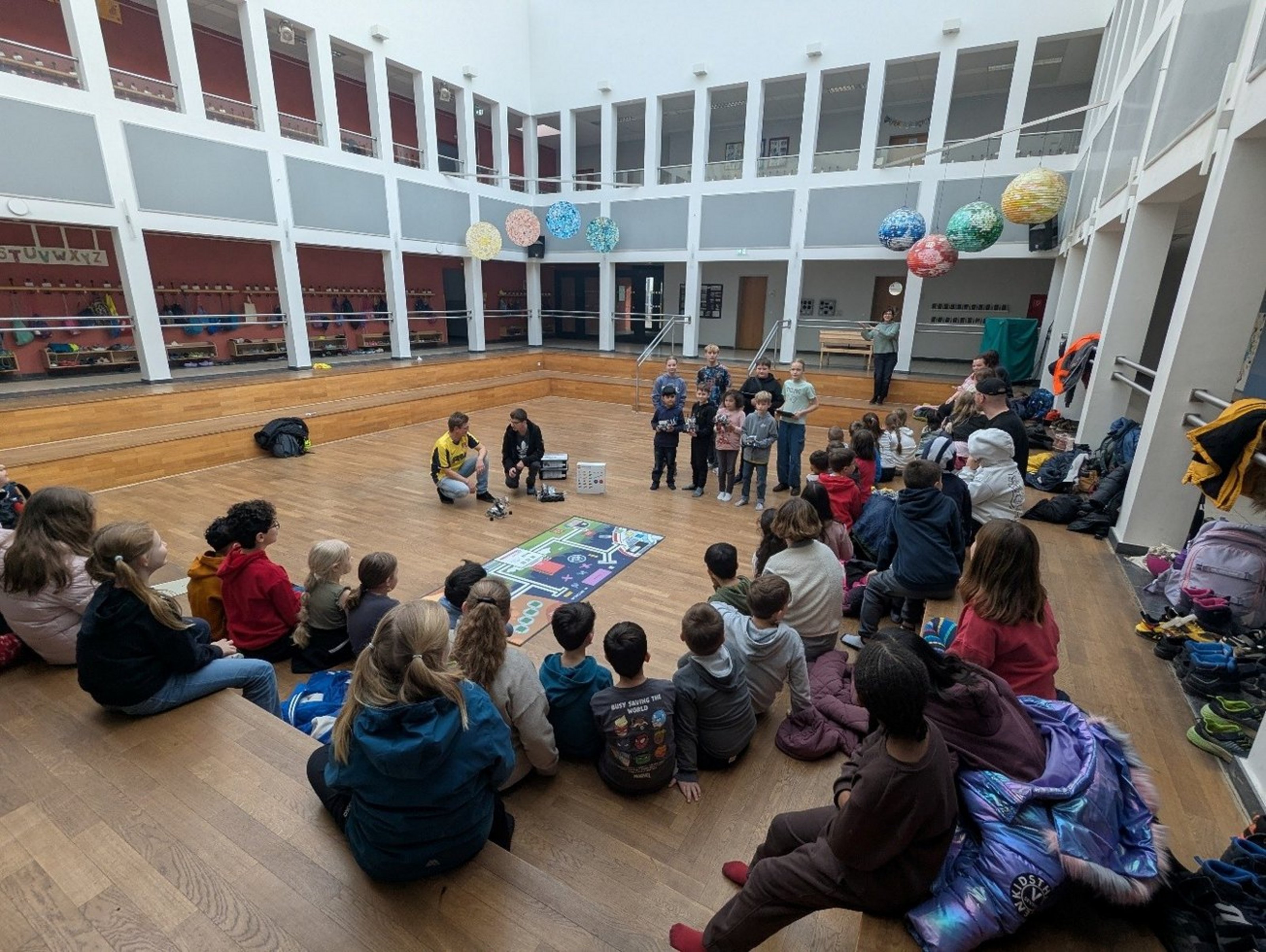 Viele Kinder sitzen und stehen in einer Runde. Zwei Kinder in der Mitte haben zwei kleine Roboter vor sich stehen. Auf dem Boden liegt ein Schaubild.
