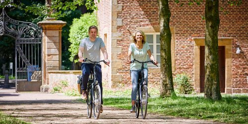 Zwei Personen fahren Fahrrad auf einem Weg, der von einem prunkvollen Tor und einem Backsteingebäude wegführt.