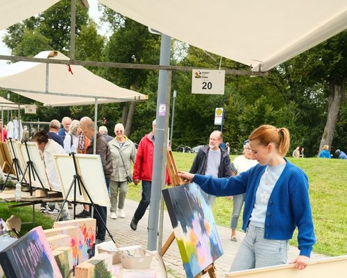 Bei einer Freiluftveranstaltung mit dem Titel „Kunst im Park“ betrachtet eine junge Frau mit rotem Pferdeschwanz und blauer Strickjacke ein farbenfrohes Gemälde auf einer Staffelei unter einem weißen Pavillon . Im Hintergrund spazieren Besucherinnen und Besucher an weiteren Ständen mit ausgestellten Kunstwerken vorbei. Die Szene spielt sich in einem grünen Parkbereich ab, es ist bewölkt aber trocken.