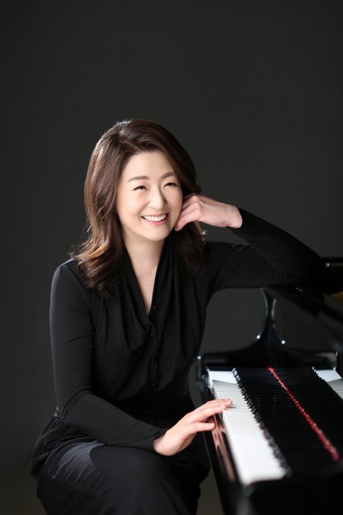 Porträt der südkoreanischen Spitzenpianistin Hee Sung Joo, lächelnd, mit langen, dunklen Haaren in schwarzer Kleidung, die an einem Flügel sitzt und sich mit einer Hand am Kopf abstützt.