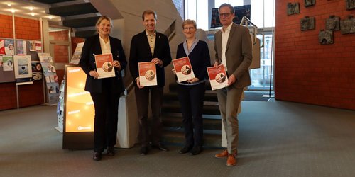 Vier Personen stehen nebeneinander an dem Fuß einer Treppe und halte jeder eine Broschüre in rot und weiß in den Händen.