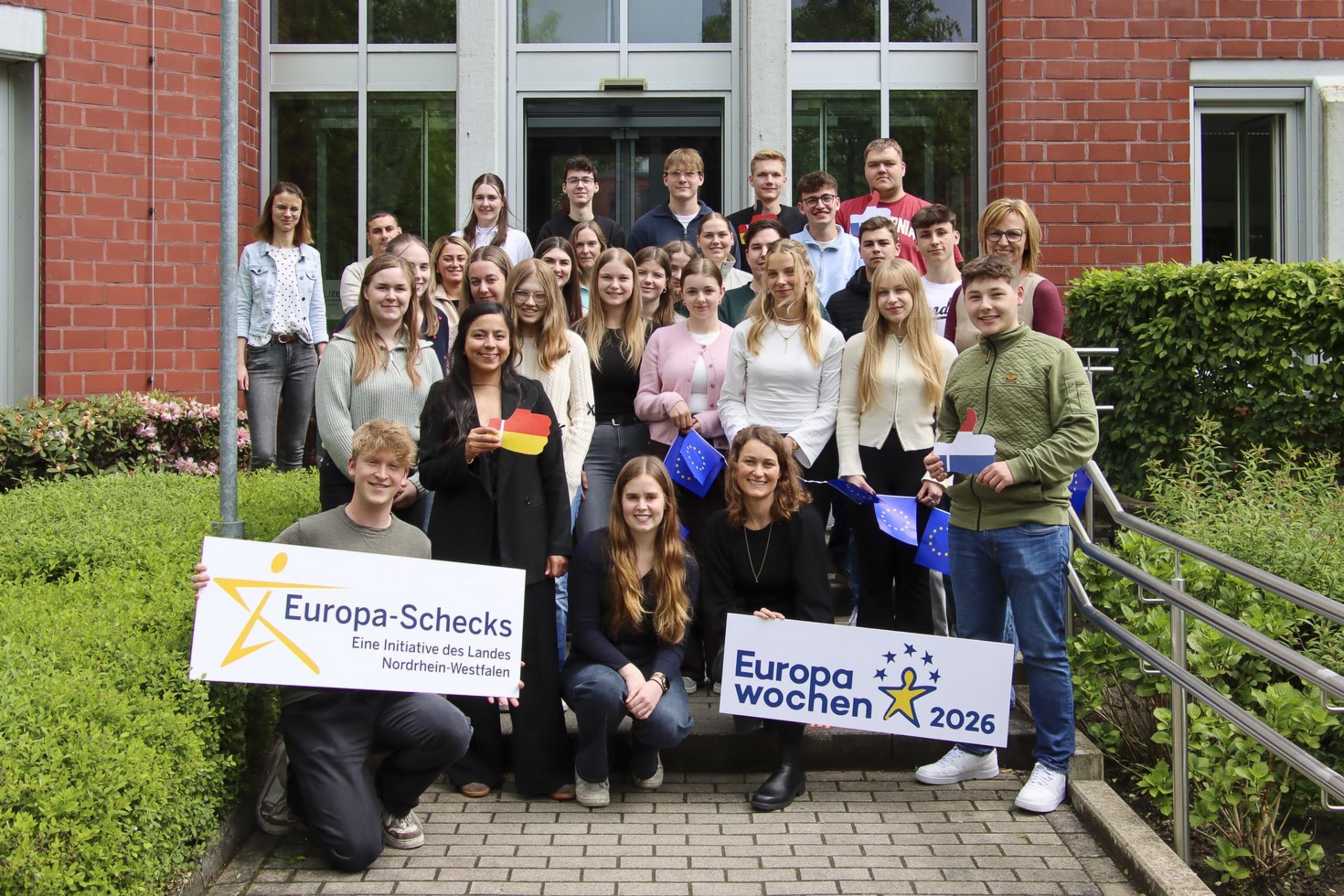 Gruppenfoto vor einem Backsteingebäude: Rund 30 Jugendliche und Erwachsene stehen auf den Stufen vor dem Eingang und blicken in die Kamera. Mehrere Personen halten Schilder mit den Aufschriften „Europa-Schecks“ und „Europawochen 2026“ sowie kleine EU-Flaggen. Die Gruppe wirkt freundlich und engagiert. Links und rechts sind Hecken und Geländer zu sehen.