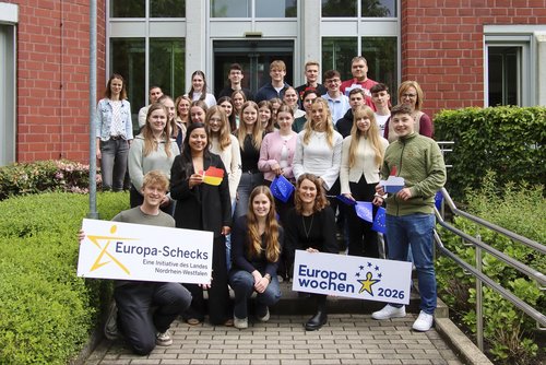 Gruppenfoto vor einem Backsteingebäude: Rund 30 Jugendliche und Erwachsene stehen auf den Stufen vor dem Eingang und blicken in die Kamera. Mehrere Personen halten Schilder mit den Aufschriften „Europa-Schecks“ und „Europawochen 2026“ sowie kleine EU-Flaggen. Die Gruppe wirkt freundlich und engagiert. Links und rechts sind Hecken und Geländer zu sehen.