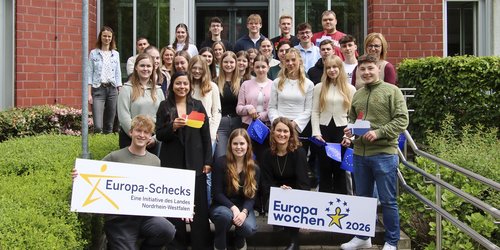 Gruppenfoto vor einem Backsteingebäude: Rund 30 Jugendliche und Erwachsene stehen auf den Stufen vor dem Eingang und blicken in die Kamera. Mehrere Personen halten Schilder mit den Aufschriften „Europa-Schecks“ und „Europawochen 2026“ sowie kleine EU-Flaggen. Die Gruppe wirkt freundlich und engagiert. Links und rechts sind Hecken und Geländer zu sehen.