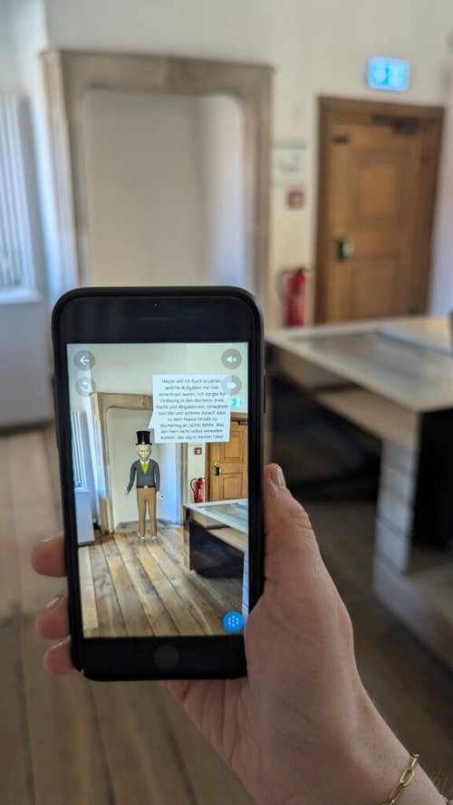 Auf einem Handy-Display ist eine Figur in einem historischen Innenraum zu sehen. 