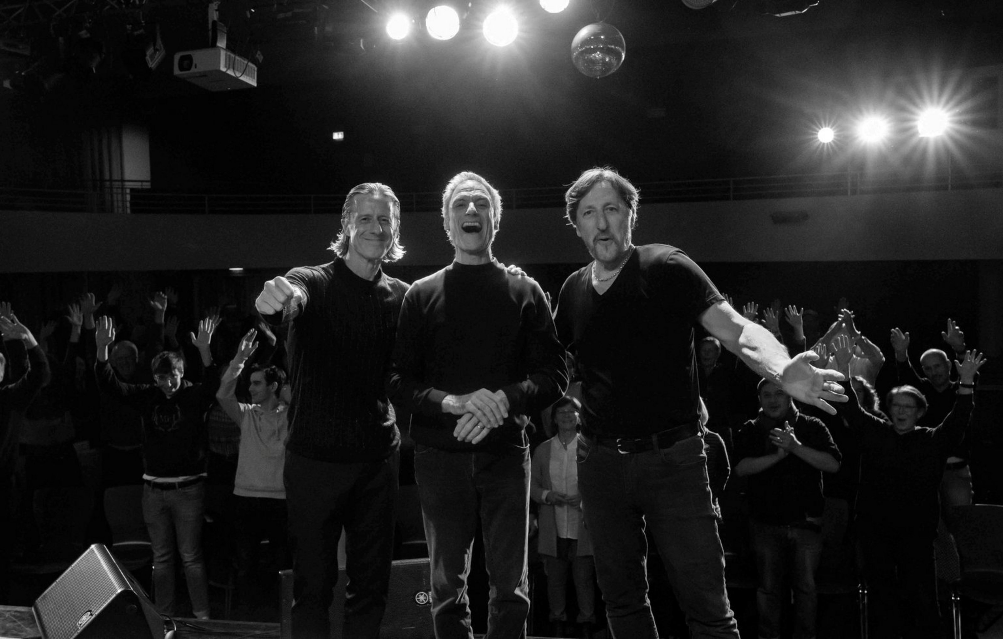 Eine Schwarz-Weiß-Aufnahme zeigt drei Männer, die nebeneinander stehen, vor ihrem Publikum in einem Saal. Alle schauen in Richtung Kamera.