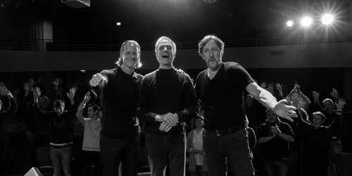Eine Schwarz-Weiß-Aufnahme zeigt drei Männer, die nebeneinander stehen, vor ihrem Publikum in einem Saal. Alle schauen in Richtung Kamera.
