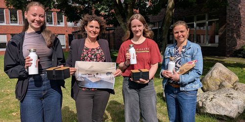 Vier Frauen stehen nebeneinander auf einer Grünfläche mit einem Baum vor einem Gebäude. Sie halten Brotdosen und Trinkflaschen und eine ein Foto von einem Klassenraum in der Hand.