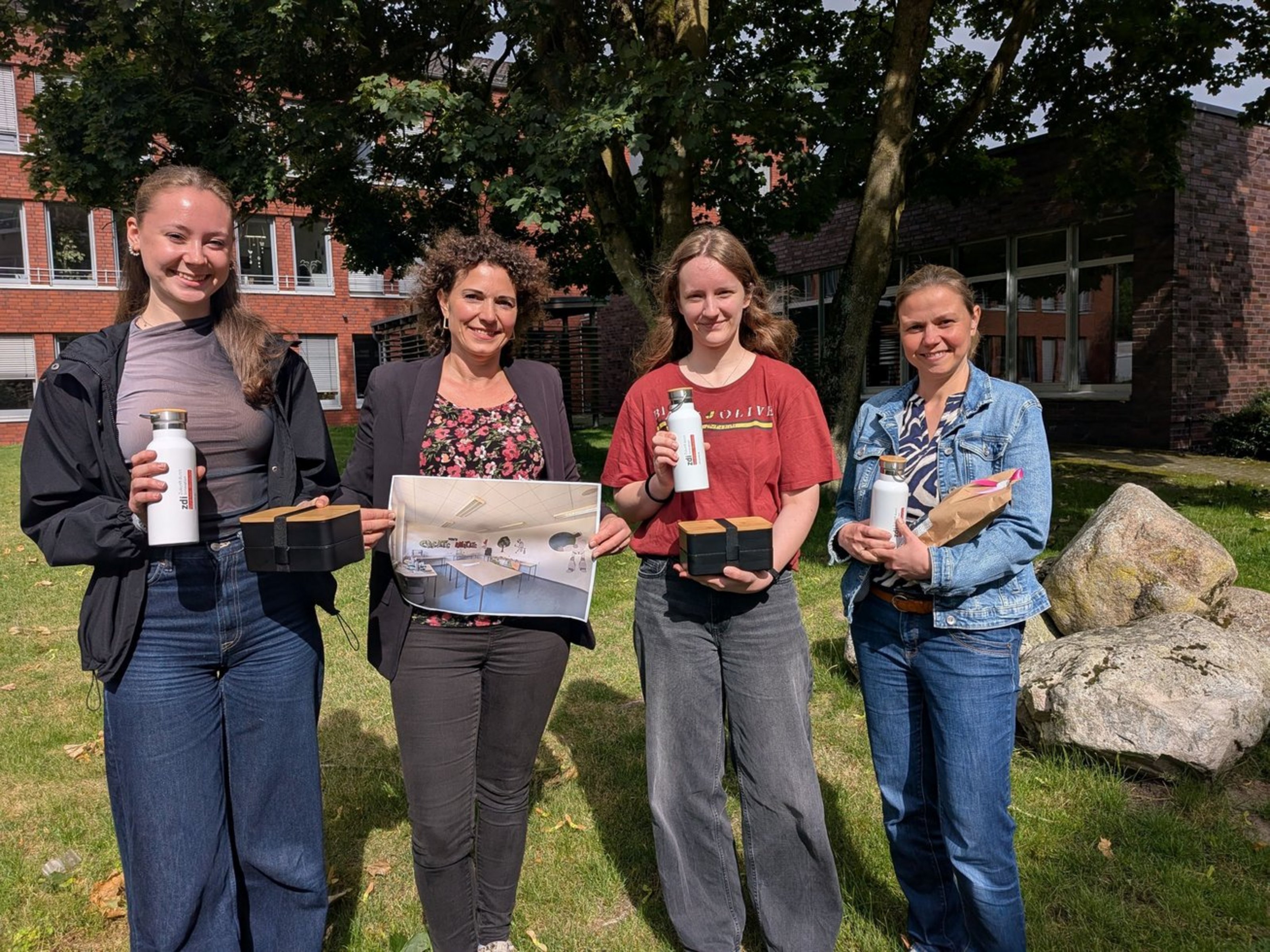 Vier Frauen stehen nebeneinander auf einer Grünfläche mit einem Baum vor einem Gebäude. Sie halten Brotdosen und Trinkflaschen und eine ein Foto von einem Klassenraum in der Hand.