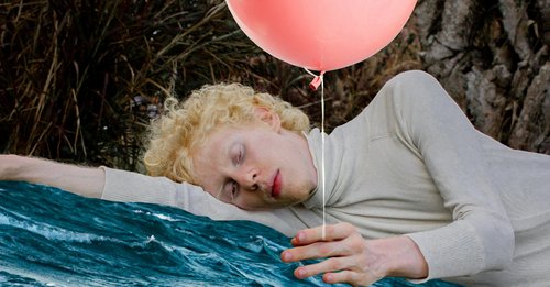 Ein Mann liegt seitlich liegend auf seinen Arm. In der anderen Hand hällt er einen rosa Luftballon. Er schaut auf seine Finger. Im Hintergrund ist Gras und ein Baumstamm zu sehen.