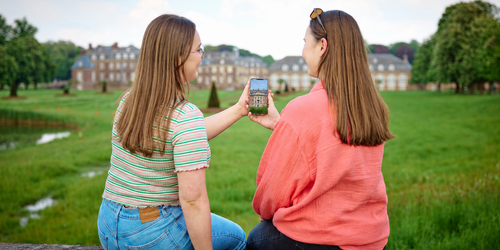 Zwei Frauen sitzen auf einer Mauer und halten gemeinsam ein Handy in der Hand. Im Hintergrund ist ein Schloss zu sehen.
