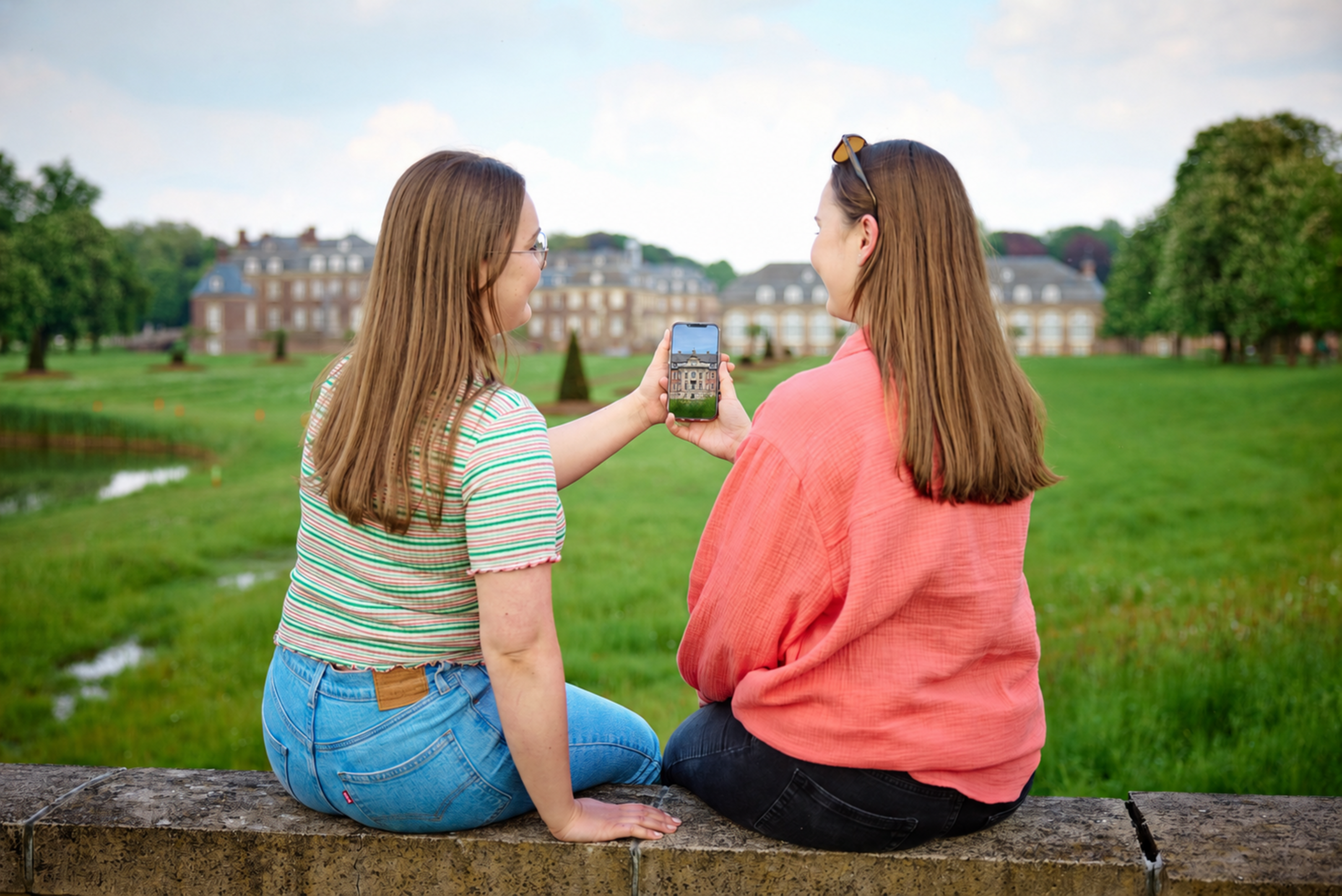 Zwei Frauen sitzen auf einer Mauer und halten gemeinsam ein Handy in der Hand. Im Hintergrund ist ein Schloss zu sehen.