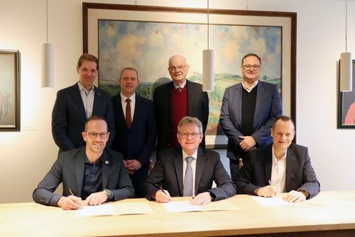 Drei Männer sitzen an einem Tisch und unterzeichnen ein Blatt Papier. Vier Männer stehen dahinter.
