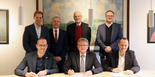 Drei Männer sitzen an einem Tisch und unterzeichnen ein Blatt Papier. Vier Männer stehen dahinter.