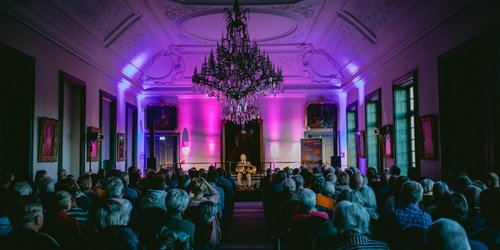 In einem Saal sitzt vor Publikum auf einer Bühne ein Mann mit Anzug und Akkordeon. Von der Decke hängt ein großer Kronenleuchter.