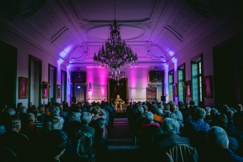 In einem Saal sitzt vor Publikum auf einer Bühne ein Mann mit Anzug und Akkordeon. Von der Decke hängt ein großer Kronenleuchter.