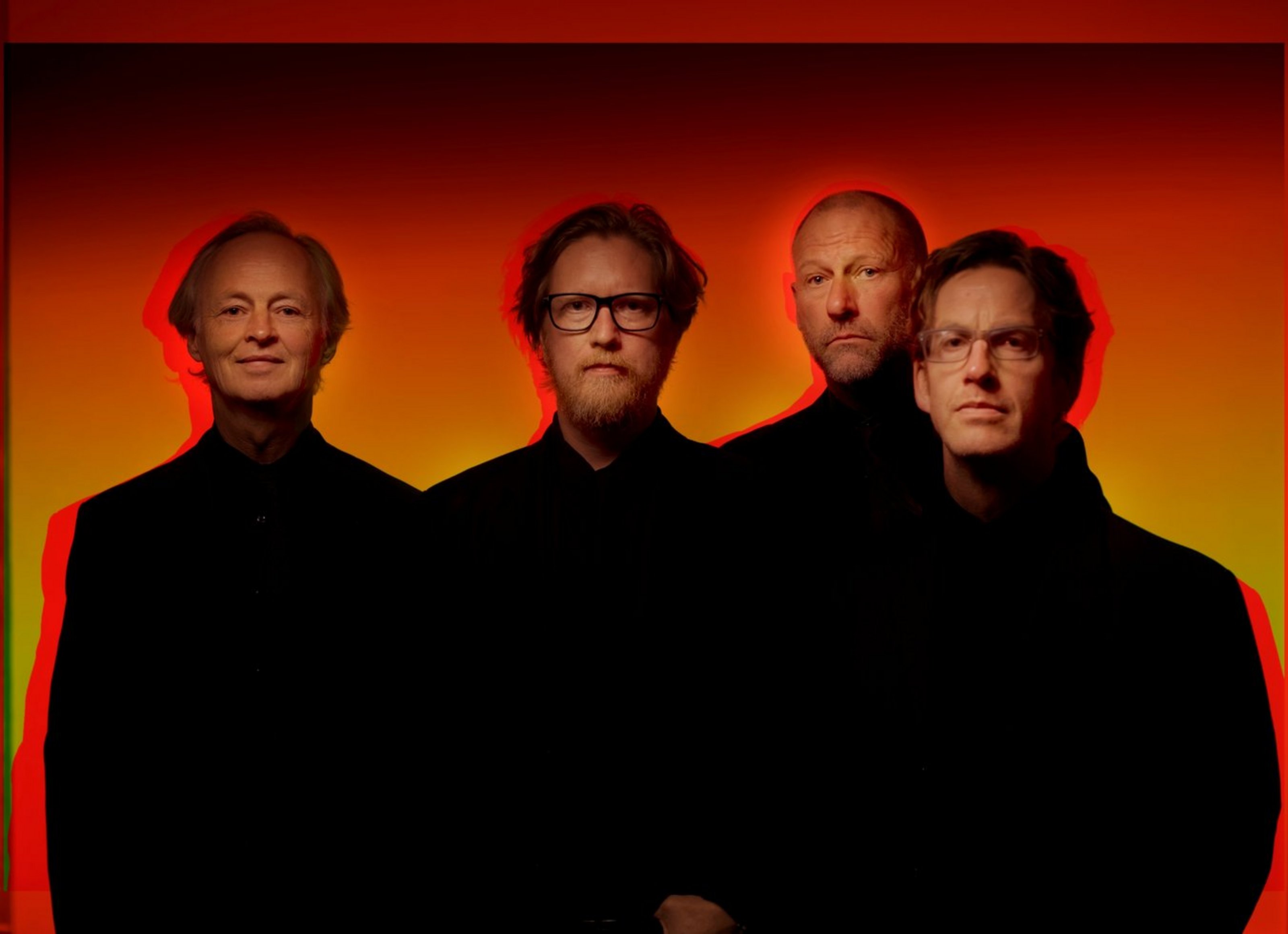 Vier Männer mit schwarzen Oberteilen stehen vor einem rot-gelbem Hintergrund und blicken in die Kamera.