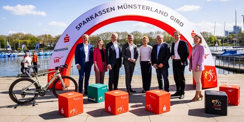 Acht Personen stehen nebeneinander vor einem Bogen mit der Aufschrift "Sparkassen Münsterland Giro". Sie stehen vor einem Steg an einem See.