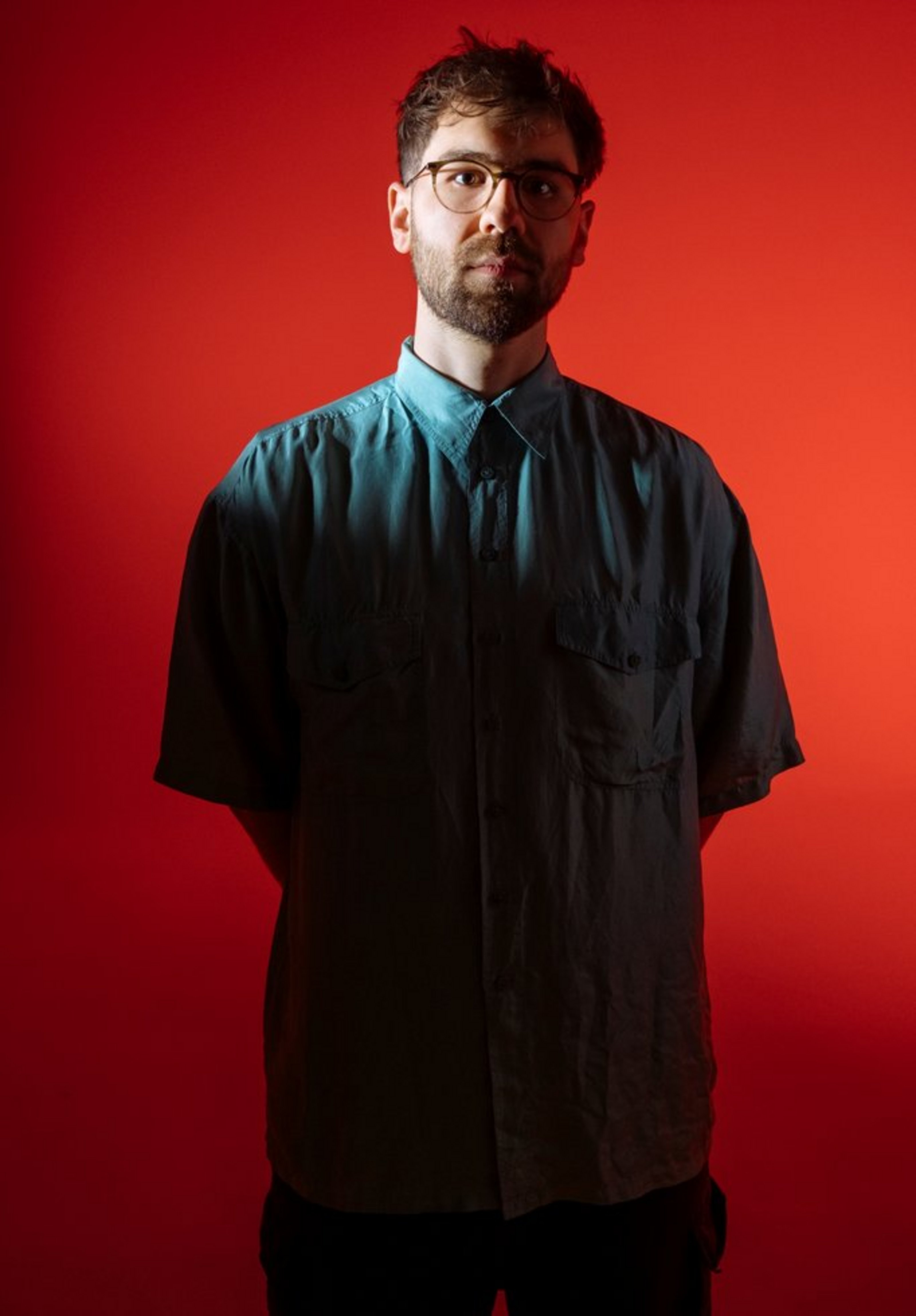 Bandleader, Tenorsaxophonist und Komponist Phillip Dornbusch (Bildquelle: Niclas Weber) Ein junger Mann mit dunklem, kurz geschnittenem Haar und Bart steht vor einem kräftig roten Hintergrund. Er trägt eine weite, kurzärmelige, petrolfarbene Hemdbluse mit zwei Brusttaschen und hat die Arme hinter dem Rücken verschränkt. Er trägt eine Brille mit runden Gläsern und blickt ruhig in die Kamera.