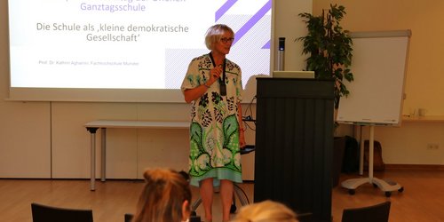 Eine Frau steht hinter einem Pult vor einer Leinwand, auf der "Selbst- und Mitentscheidungsräume eröffnen: Partizipation im Alltag der Offenen Ganztagsschule. Die Schule als 'kleine demokratische Gesellschaft'".