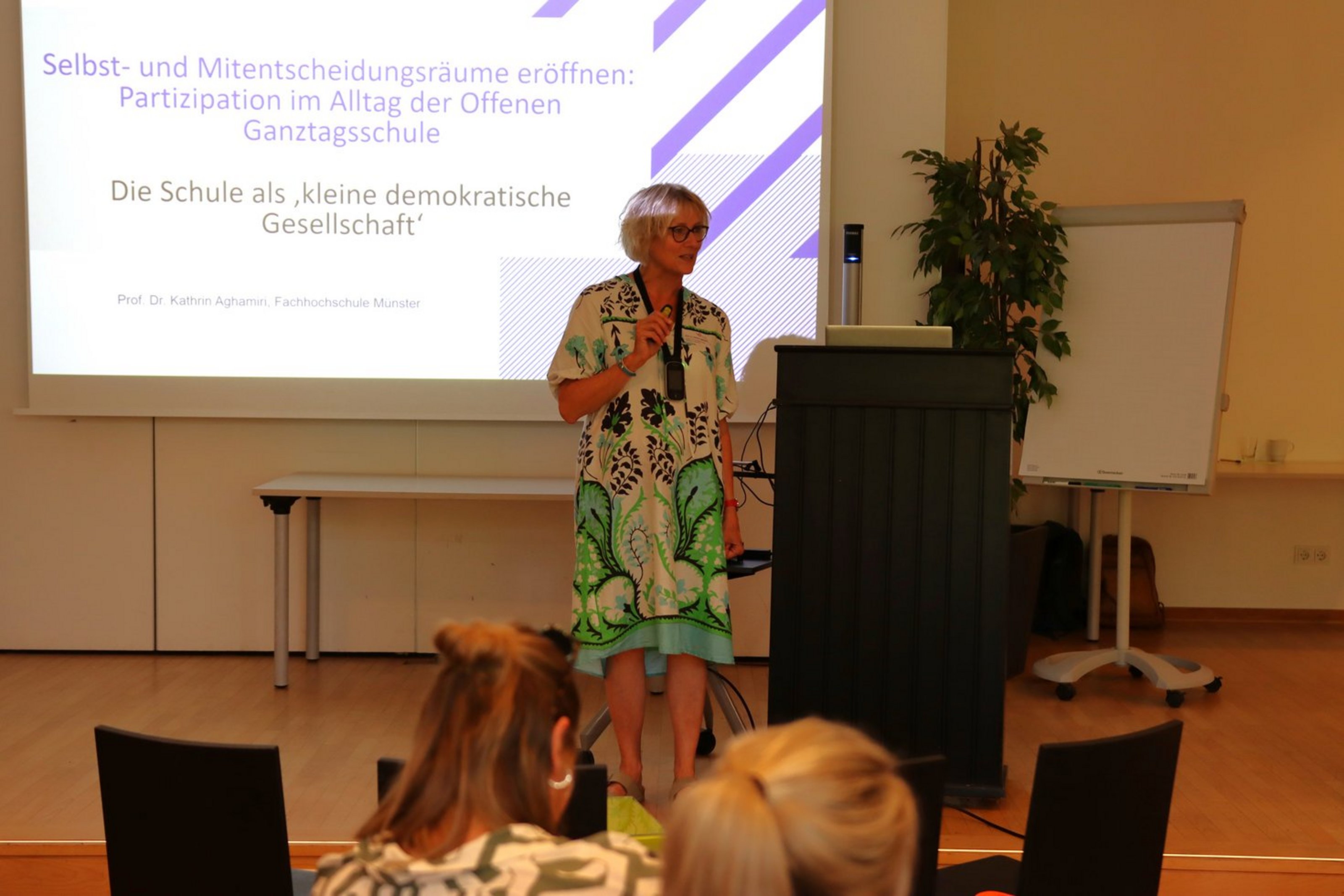 Eine Frau steht hinter einem Pult vor einer Leinwand, auf der "Selbst- und Mitentscheidungsräume eröffnen: Partizipation im Alltag der Offenen Ganztagsschule. Die Schule als 'kleine demokratische Gesellschaft'".