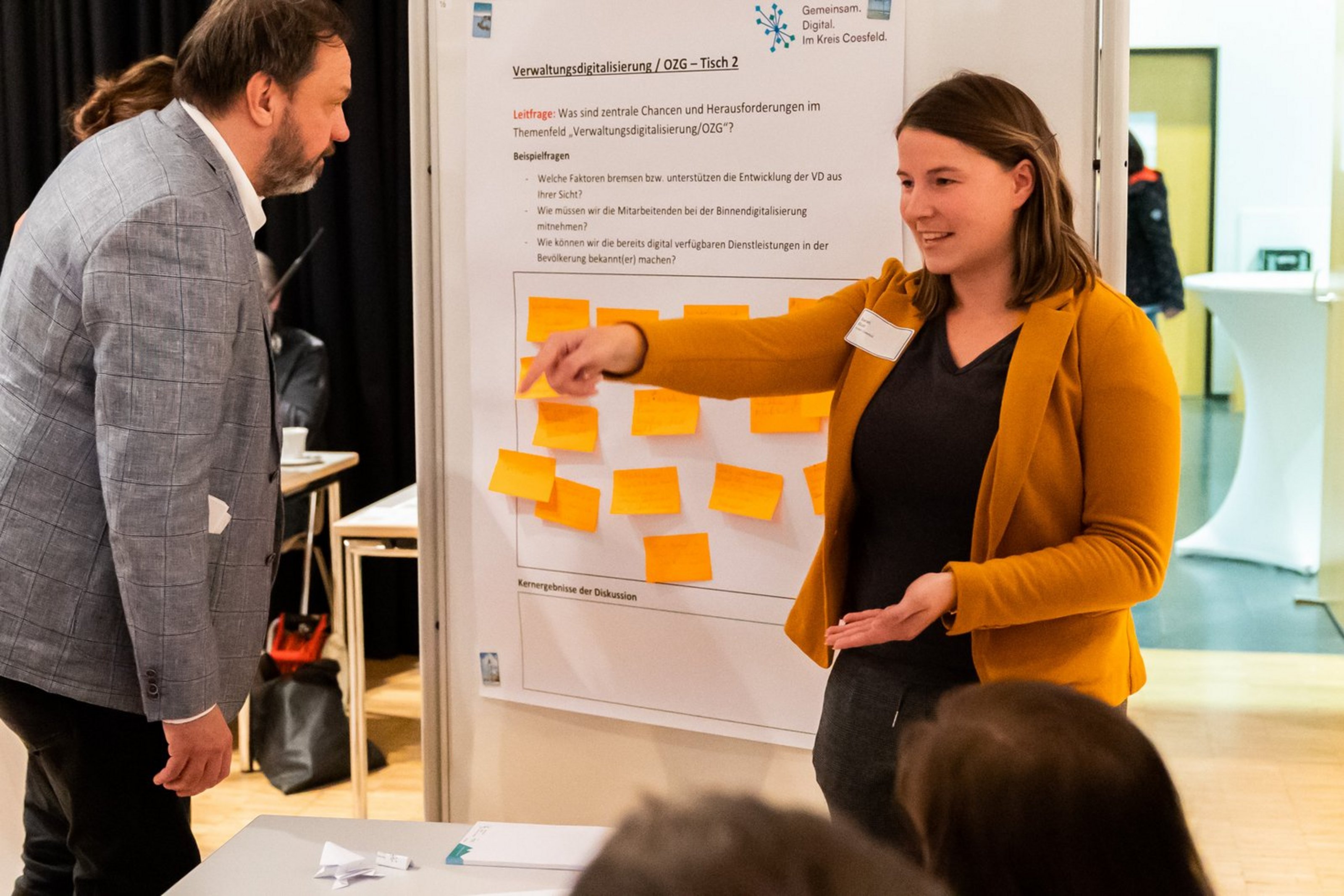Frau in senfgelbem Blazer erklärt einem Mann vor einem Flipchart mit vielen gelben Haftnotizen etwas