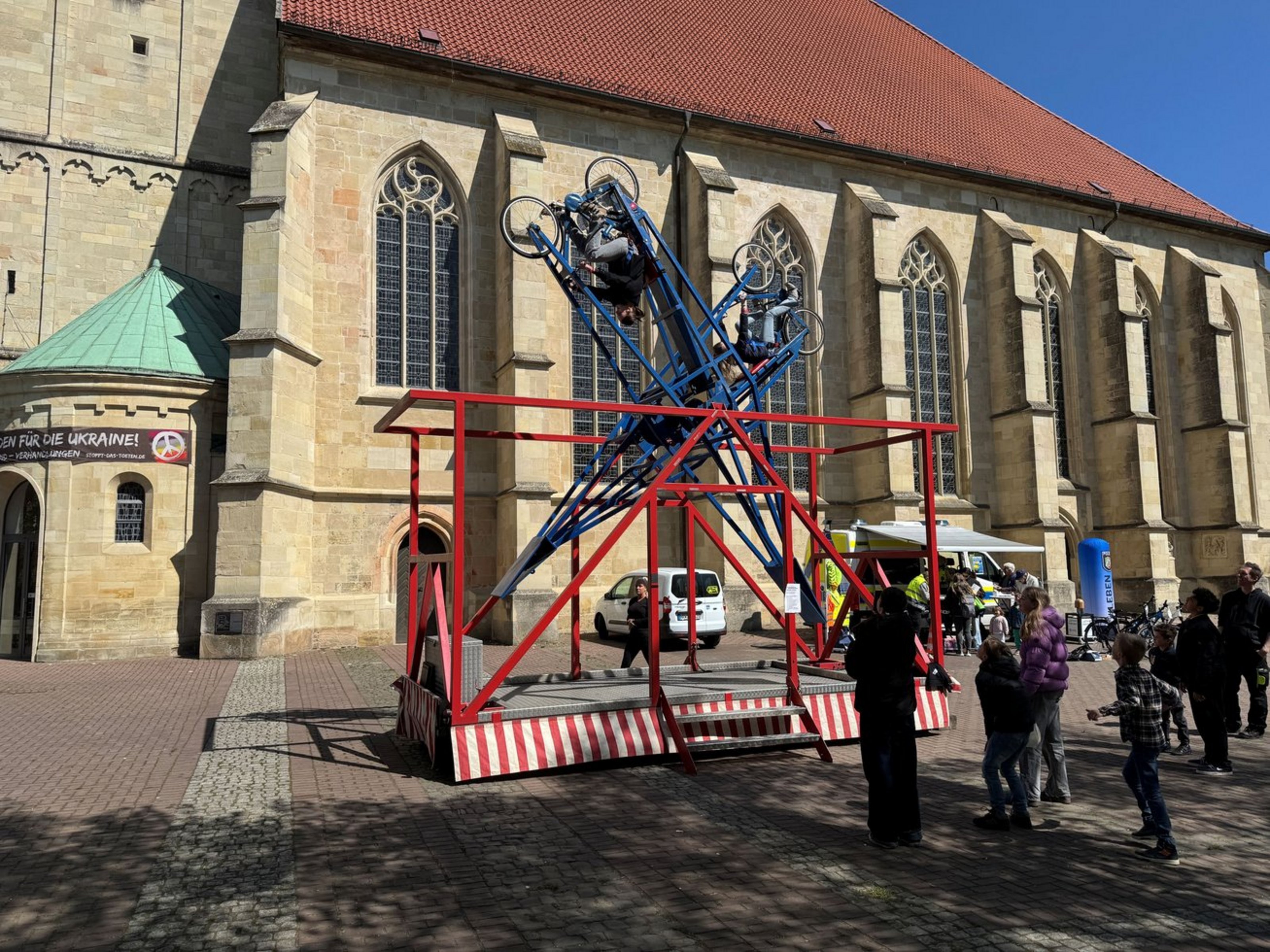 Vor einer Kirche steht ein Karussel, in das zwei Personen mit Fahrrad eingespannt sind. Sechs Personen schauen zu.