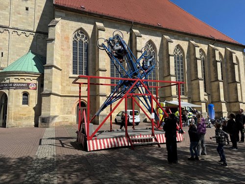 Vor einer Kirche steht ein Karussel, in das zwei Personen mit Fahrrad eingespannt sind. Sechs Personen schauen zu.