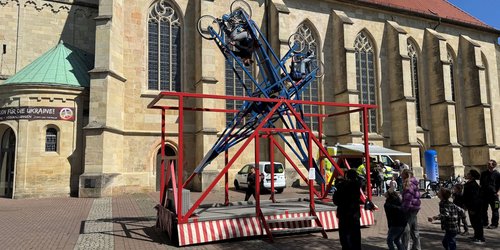Vor einer Kirche steht ein Karussel, in das zwei Personen mit Fahrrad eingespannt sind. Sechs Personen schauen zu.