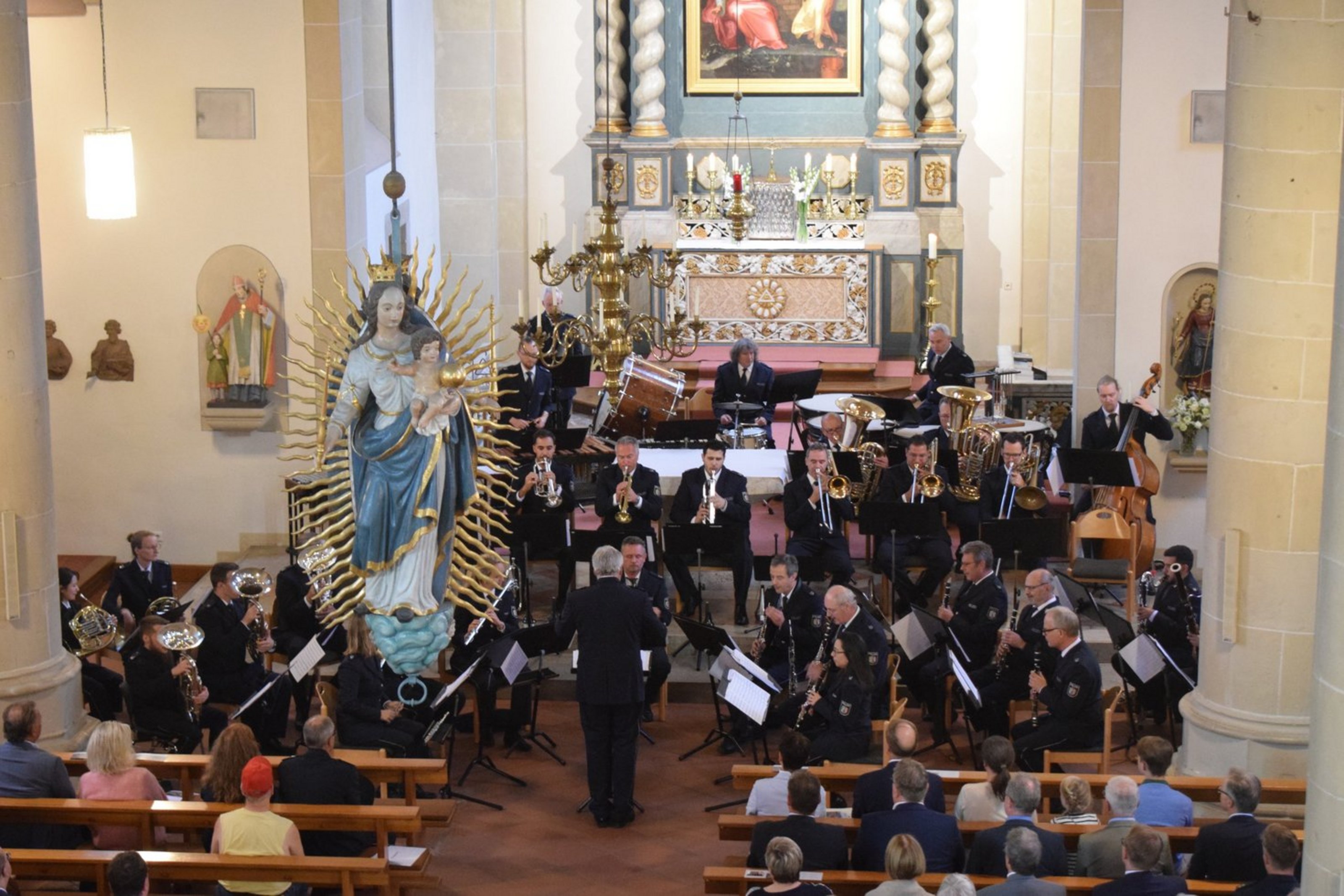 Das Landespolizeiorchester spielt in Ascheberg (Fotoquelle: Kreis Coesfeld). Ein Blasorchester tritt in einer Kirche auf. Die Musikerinnen und Musiker sitzen in mehreren Reihen vor dem Altar, während ein Dirigent vor ihnen steht. Die Kirche ist mit religiösen Statuen und einem großen Marienbildnis mit Jesuskind geschmückt, das prominent im Vordergrund links hängt. Im Hintergrund sind der Altar, ein Gemälde und ein Kronleuchter zu sehen. Die Zuschauer sitzen in Kirchenbänken und verfolgen das Konzert.