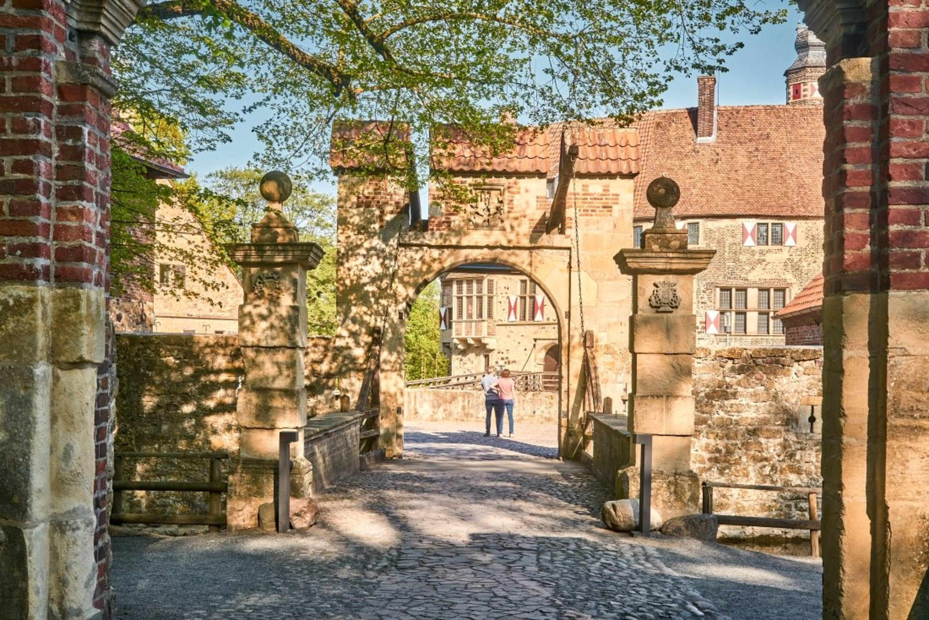 Auf dem Innnenhof einer Burg stehen zwei Personen. Der Zugang zum Innenhof führt über eine Brücke mit zwei Toren. Im Hintergrund steht ein großes historisches Backsteingebäude. Der Himmel ist blau.