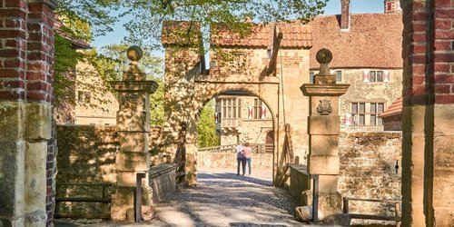Auf dem Innnenhof einer Burg stehen zwei Personen. Der Zugang zum Innenhof führt über eine Brücke mit zwei Toren. Im Hintergrund steht ein großes historisches Backsteingebäude. Der Himmel ist blau.