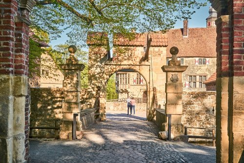 Auf dem Innnenhof einer Burg stehen zwei Personen. Der Zugang zum Innenhof führt über eine Brücke mit zwei Toren. Im Hintergrund steht ein großes historisches Backsteingebäude. Der Himmel ist blau.