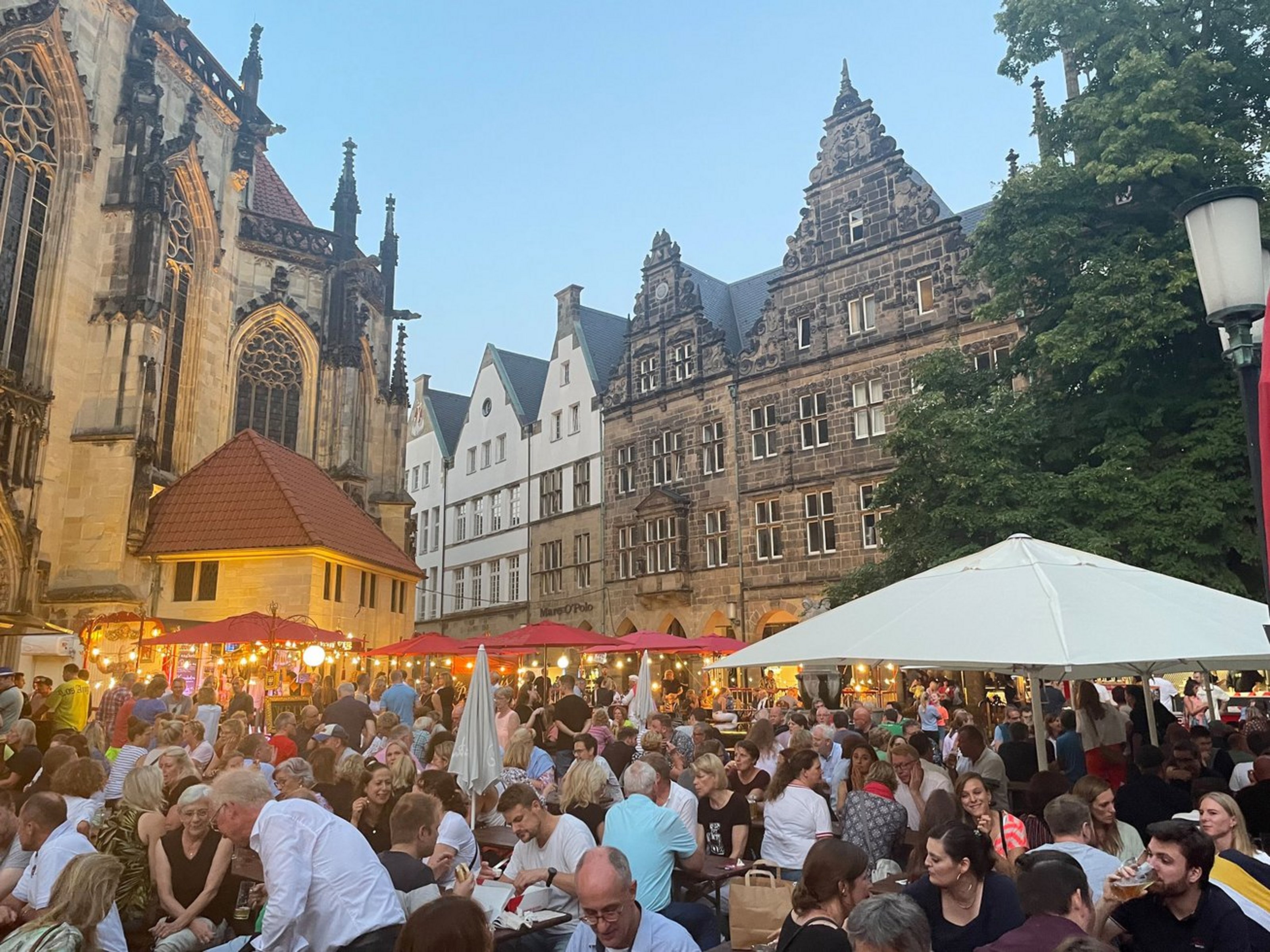 Man sieht eine große Menga Menschen abends zusammen auf einem Marktplatz sitzen.