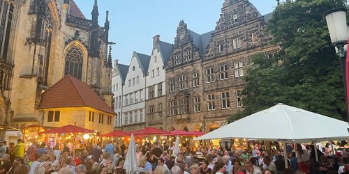 Man sieht eine große Menga Menschen abends zusammen auf einem Marktplatz sitzen.