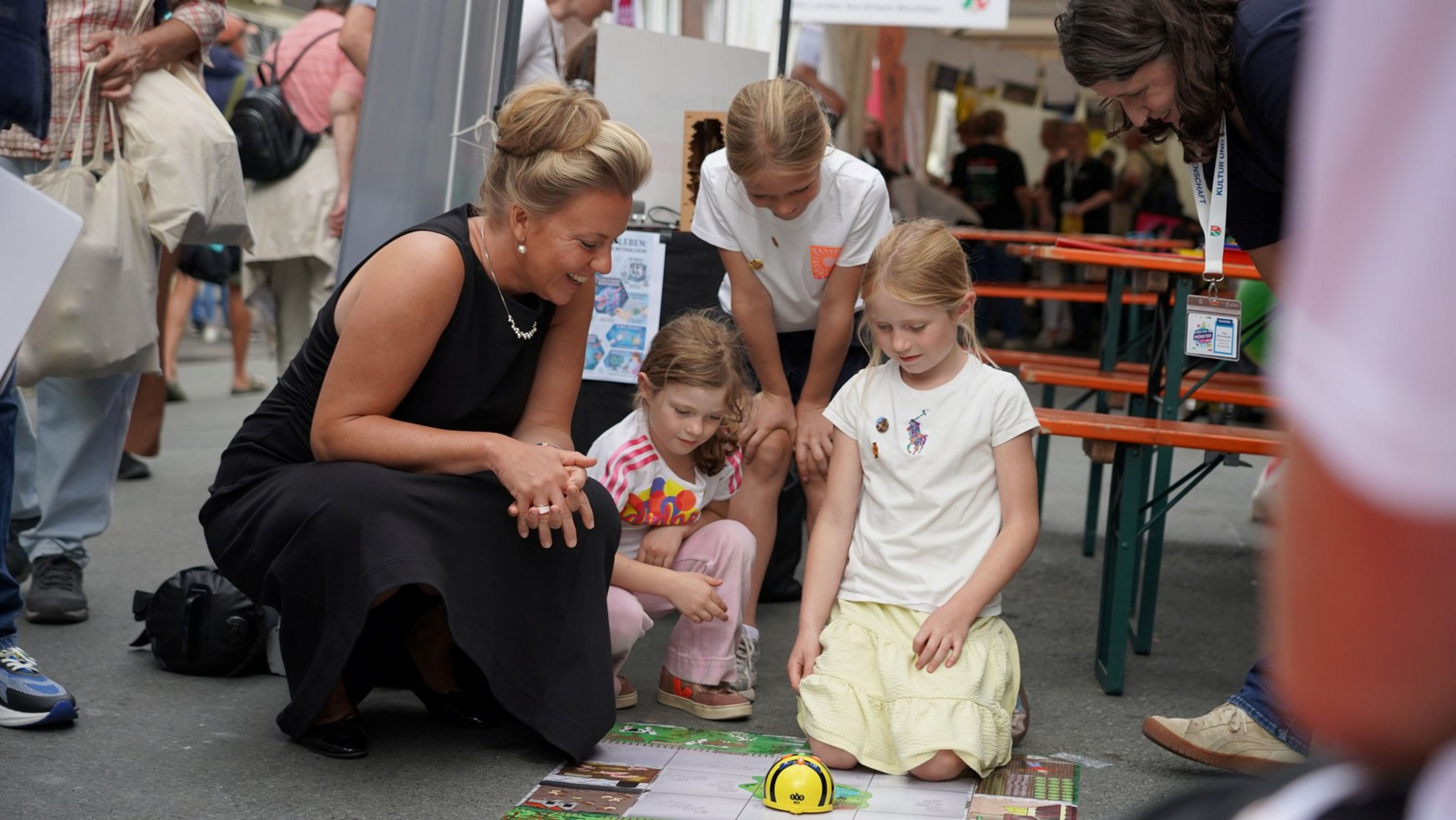 Eine Frau und drei Kinder knien auf einer Straße und schauen einen bunten Gegenstand auf dem Boden an. Im Hintergrund stehen Holzbänke und weitere Menschen.