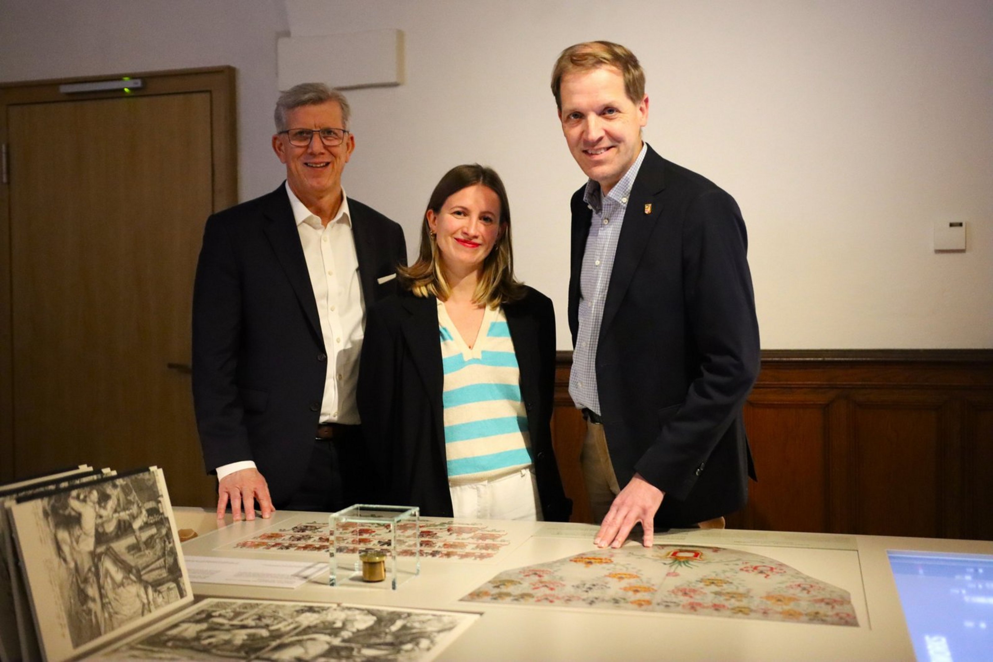 Drei Personen stehen in der Ausstellung der Burg Vischering und präsentieren historische Exponate, darunter ein Petschaftsiegel, das auf einem Tisch vor ihnen liegt.