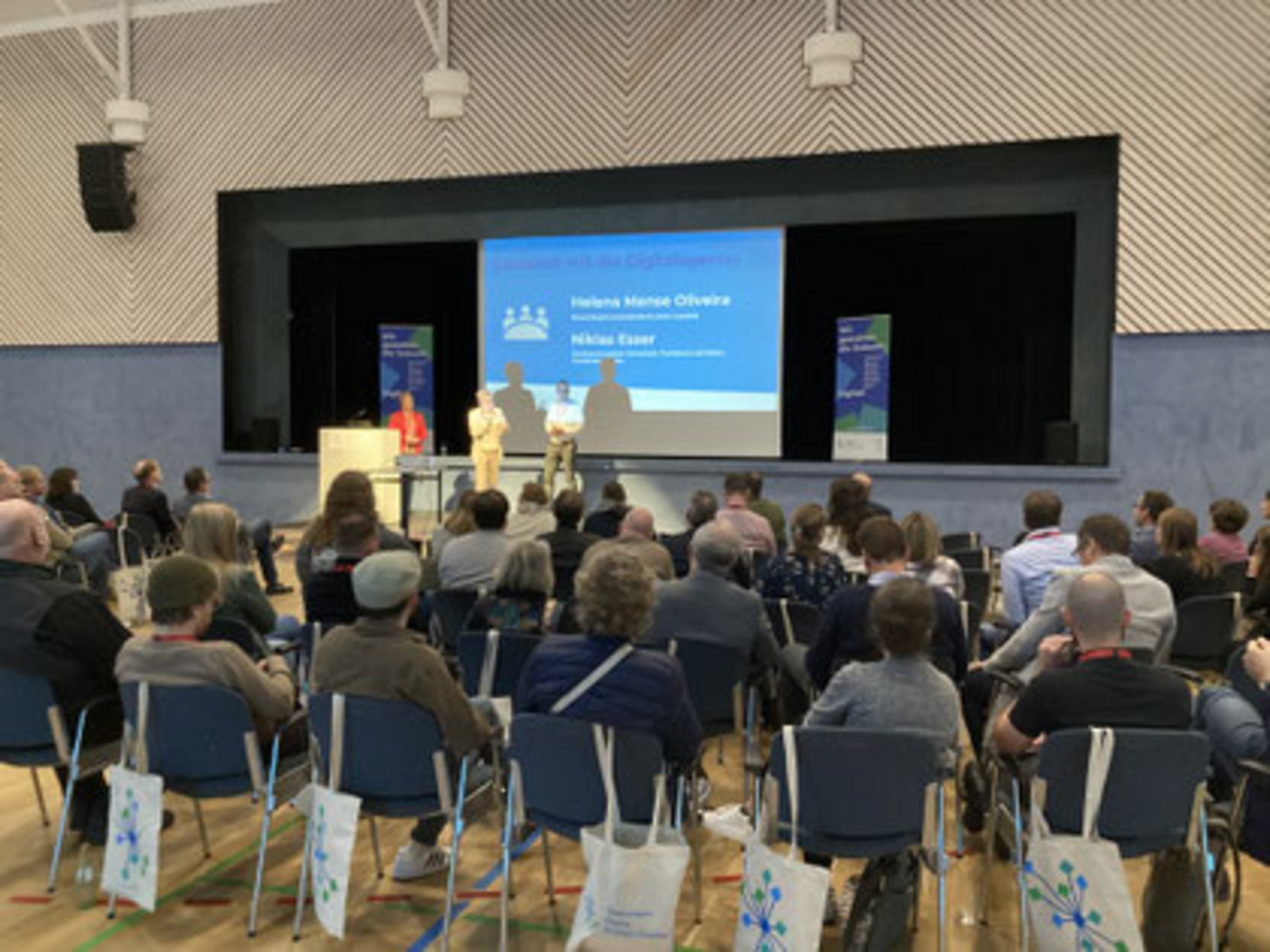 Mehrere Personen sitzen in einem Saal und blicken auf eine Bühne, auf der drei Menschen vor einer großen Leinwand stehen.