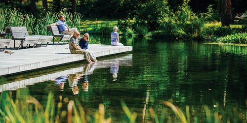 Auf einem Steg an einem See sitzen vier Personen. drei halten ihre Füße ins Wasser. Im Hintergrund sind Bäume zu sehen.